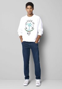Weißer Sweatshirt mit Grafikdruck, der den Text "CASA WAY" und eine Figur zeigt, kombiniert mit dunkelblauen Jeans und weißen Sneakern.