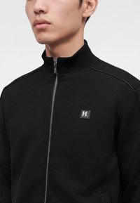 Jeune homme portant une veste noire à zip avec un col montant et un petit logo argenté sur la poitrine.