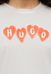 Ανοιχτό γκρι μπλουζάκι με τη λέξη "HUGO" σε πορτοκαλί καρδιές, με μαλακή υφή και στρογγυλή λαιμόκοψη.