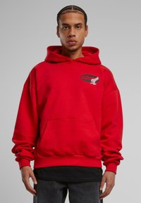 Giovane con capelli intrecciati che indossa una felpa rossa con un logo nero sul petto e pantaloni scuri, in piedi su uno sfondo grigio uniforme.