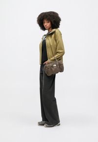 Αδιάβροχο μπουφάν ελιάς πάνω από ένα μαύρο τοπ, σετ με σκούρο παντελόνι φαρδιάς γραμμής και καφέ τσάντα duffel με απαλή υφή. Παπούτσια με καουτσούκ σόλα.