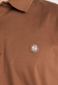 Barna póló, puha textúrával, gallérral és egy kör alakú logo folttal, amely ezüst színű "BB" betűket ábrázol.