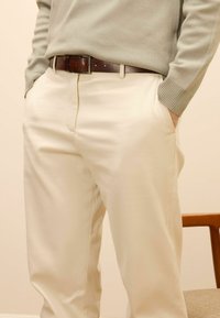 Beige broek met een gladde structuur, iets taps toelopende pijpen en zijzakken, gecombineerd met een grijze trui en een bruin leren riem.