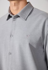 Camicia grigia chiaro con colletto, realizzata in tessuto testurizzato. Presenta un piccolo logo di un uccello ricamato nella zona sinistra del petto.