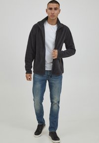 Mørk grå fleece-jakke med glidelås og høy krage, brukt over en hvit t-skjorte. Tettsittende design kombinert med lyseblå jeans og svarte joggesko.