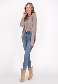 Pulover tricotat maro deschis, scurt, blugi cu talie înaltă de culoare albastră și cizme de gleznă în stil vest, de nuanță bej, cu detalii perforate. Fundal alb.