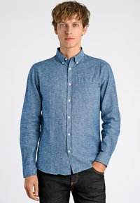 Heren langemouwen button-up shirt in lichtblauwe chambray stof, met een borstzak en witte knopen, gecombineerd met donkere jeans.