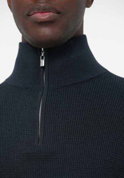 Pulover tricotat de culoare bleu închis, cu guler înalt și închidere cu fermoar pe jumătate, având o suprafață texturată și detalii cu fermoar neted.