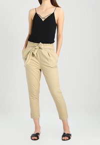 Svart ärmlös topp med korskorsade axelband, matchad med ljusbeige tapered-byxor med bälte och elastisk midja. Svarta slidesandaler.