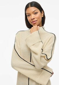 Pull en maille beige avec un col montant, présentant des finitions noires le long des coutures et des manches larges, mettant en valeur un tissu texturé.