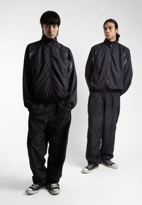 Black windbreaker tracksuits with a textured pattern and contrasting gray accents, paired with black sneakers, worn by two models.