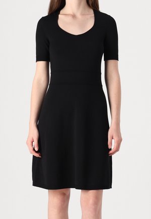Femme portant une robe noire jusqu'au genou avec manches courtes et décolleté en V, debout devant un fond clair uni.
