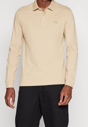 Beżowa koszulka polo z długim rękawem, z kołnierzykiem i haftowanym logo na piersi, zestawiona z ciemnymi szortami. Gładka tkanina bawełniana.