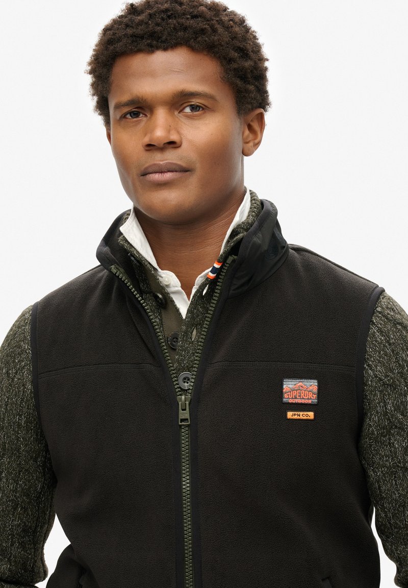 Gilet en polaire noir avec fermeture éclair, présentant un logo en patch, superposé à une chemise verte texturée avec col et boutons.
