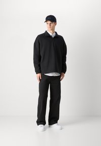 Felpa nera con zip a metà, vestibilità rilassata, abbinata a una camicia bianca con colletto. Pantaloni neri e sneaker bianche completano l'outfit. Cappellino nero in accento.