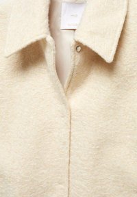 Beige, texturovaná bunda s límcem, vybavená jedním cvokem a měkkou, huňatou tkaninou. Viditelná vnitřní etiketa.