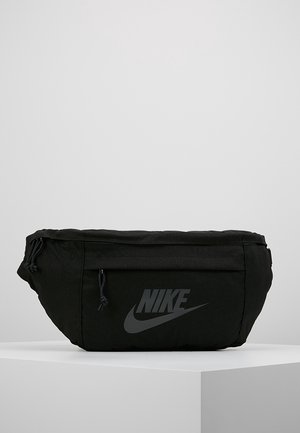 Sac banane noir Nike avec poche avant zippée et logo de marque, placé sur un bloc blanc sur fond gris uni.