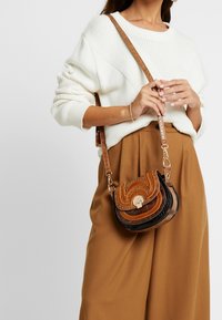 Brązowa skórzana torba na ramię z teksturowanymi akcentami, zaokrąglonymi kształtami i złotym zapięciem. Nosi ze swetrem w kolorze kremowym i brązowymi spodniami.
