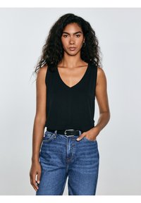 Vrouw met lang krullend haar die een mouwloze zwarte V-hals top draagt, ingestopt in een high-waisted blauwe spijkerbroek met een zwarte riem.