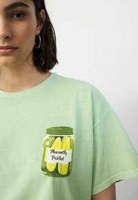 T-shirt vert menthe avec un motif d'un bocal avec un couvercle vert et des cornichons jaunes, étiqueté "Agrémenté avec plaisir". Matière en coton doux.