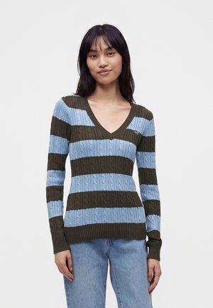 Jeune femme portant un pull col en V avec de larges rayures horizontales bleues et vert foncé ainsi qu'un jean bleu clair, debout devant un fond uni.
