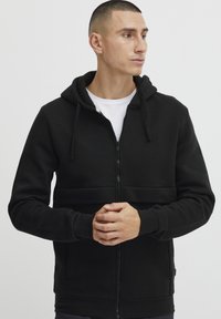 Sudadera negra con cierre de cremallera y capucha ajustable con cordones, cierre frontal de cremallera y bolsillos tipo canguro. Tejido suave y texturizado con un diseño ajustado.