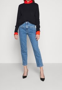 Mujer vestida con un suéter negro sobre una camisa roja con cuello, jeans de tiro alto azul y tacones de slingback negros con punta, de pie contra una pared lisa.