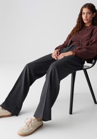 Femme avec de longs cheveux bouclés, portant une veste marron et un large pantalon gris, assise sur une chaise noire contre un fond clair et unicolore.