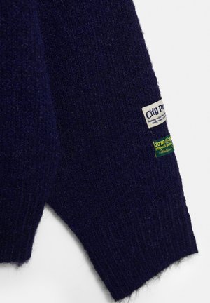 Dunkelblauer Strickpulloverärmel mit zwei kleinen Stoffetiketten, eines weiß mit Text und eines grün mit gelbem Text.