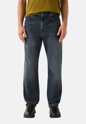 Mann trägt lockere dunkelblaue Jeans, schwarze Sneakers und ein senfgelbes Hemd, steht mit entspannten Händen an den Seiten.