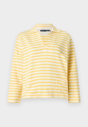 Kobieta stojąca, ubrana w żółto-biały, paskowany top z długim rękawem, niebieskie dżinsy, białe trampki, trzyma brązową torebkę w lewej ręce.