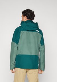 Veste imperméable en bleu sarcelle foncé et vert clair, avec capuche, panneaux contrastants et logo dans le haut du dos. Tissu texturé avec finition lisse.
