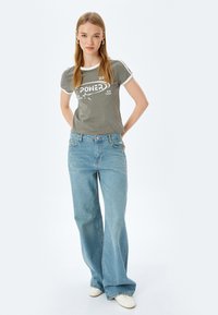 Lichtgrijze t-shirt met witte afwerking, voorzien van een grafisch ontwerp. Gepaard met lichtblauwe wijde jeans en witte sneakers. Eenvoudige achtergrond.