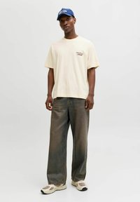 Μπες κοντομάνικο μπλουζάκι σε μπεζ απόχρωση με εκτύπωση "Origins", συνδυασμένο με σκούρο γκρι παντελόνι με φαρδιά γραμμή και κρέμ αθλητικά παπούτσια. Το λουκ ολοκληρώνει ένα ναυτικό καπέλο.