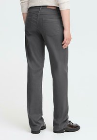 Person med mørkegrå rette bukser, beige genser og svarte loafers, som står med ryggen til kameraet mot en ensfarget lys bakgrunn.