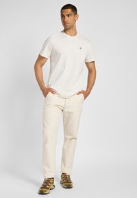 T-shirt crema a maniche corte con un piccolo logo blu, abbinato a pantaloni leggeri beige e sneakers marroni multicolore con un design in rete.
