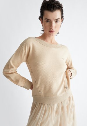 Maglione beige a maglia con motivo testurizzato, scollo a barca oversize, dettagli con bottoni ai lati e polsini a costine. Gonna in tulle trasparente sottostante.