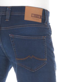 Jeans dżinsowe w ciemnoniebieskim odcieniu, z pomarańczowym przeszyciem i brązową skórzaną metką oznaczoną "MUSTANG True Denim" na tylnej części pasa.