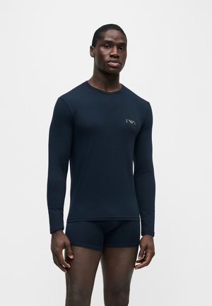 Mann trägt eng anliegendes langärmeliges navyfarbenes Hemd mit kleinem goldenem Logo und passende navyfarbene Boxershorts vor einfarbig weißem Hintergrund.