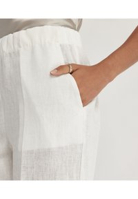Weiße Leinenhose mit strukturiertem Finish, ausgestattet mit einem elastischen Bund und seitlichen Taschen. Eine Hand ruht in der Tasche.