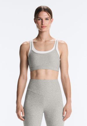 Jeune femme aux cheveux bruns attachés, portant un soutien-gorge de sport gris avec un liseré blanc et des leggings taille haute assortis, debout face à l'avant.