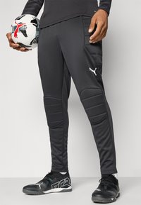 Čierne športové nohavice s polstrovanými kolenami, zúžený strih a bielym logom. Model drží čierno-biely futbalový loptu a má na sebe čierne športové topánky.
