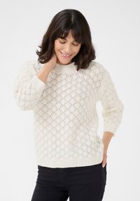 Pull en maille crème avec un motif texturé et ajouré, encolure ronde et poignets côtelés. Associé à un pantalon sombre, mettant en avant une coupe décontractée.