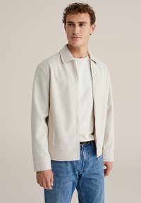 Chaqueta beige claro con cremallera, hecha de tela suave, con cuello plano, bolsillos laterales y puños ajustados, combinada con vaqueros azules.