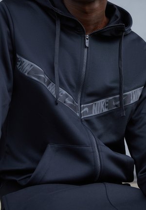 Black athletic zip-up hoodie with a smooth texture, featuring a prominent gray Nike logo stripe across the chest and a kangaroo pocket.