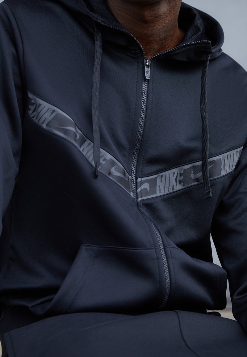 Černá sportovní mikina na zip s hladkou texturou, vyznačující se výrazným šedým pruhem s logem Nike přes hrudník a klokankovou kapsou.