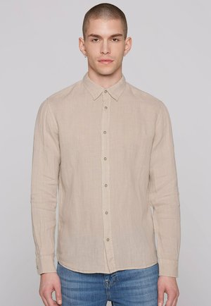 Junger Mann mit kurzen Haaren, der ein beiges, langärmliges Hemd mit Knöpfen und blaue Jeans trägt, steht vor einem hellgrauen Hintergrund.