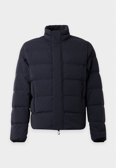 Veste matelassée bleu marine foncé avec col haut, fermeture éclair frontale dissimulée et poches latérales zippées, affichant un petit logo sur la poitrine gauche.
