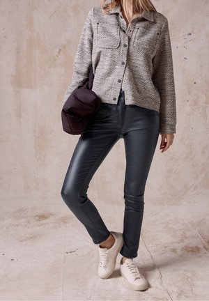 Kvinde iført grå tekstureret skjorte med knapper, sorte læderbukser, hvide sneakers, der holder en mørk dynejakke foran en beige tekstureret baggrund.