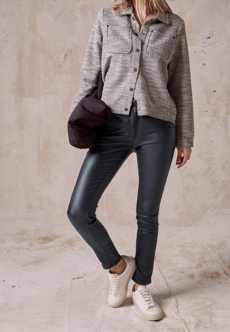 Femme portant une chemise grise texturée à boutons, un pantalon en cuir noir, des baskets blanches, tenant une doudoune sombre devant un fond beige texturé.